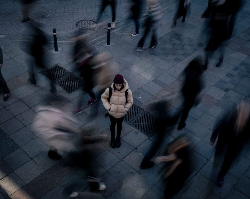 A woman standing still on a city sidewalk as blurred people walk around her, symbolizing isolation amidst u...
