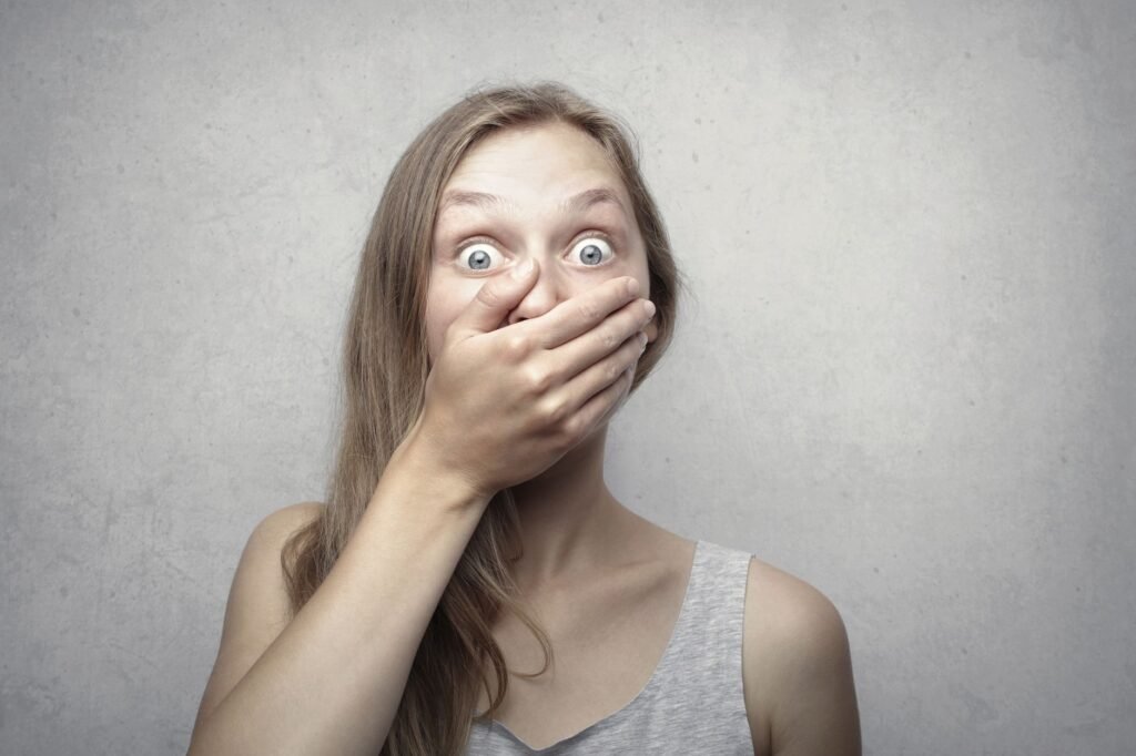 Wide eyed woman with long hair covering her mouth with one hand against a gray wall