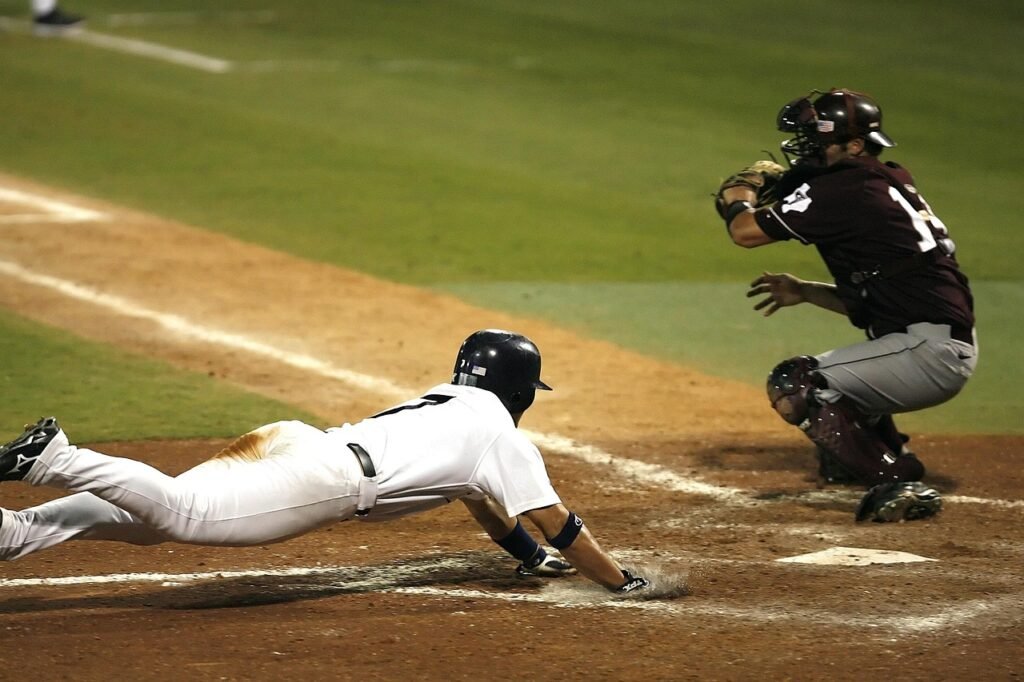Baseball player diving headfirst toward the plate as the catcher waits nearby on a dirt infield