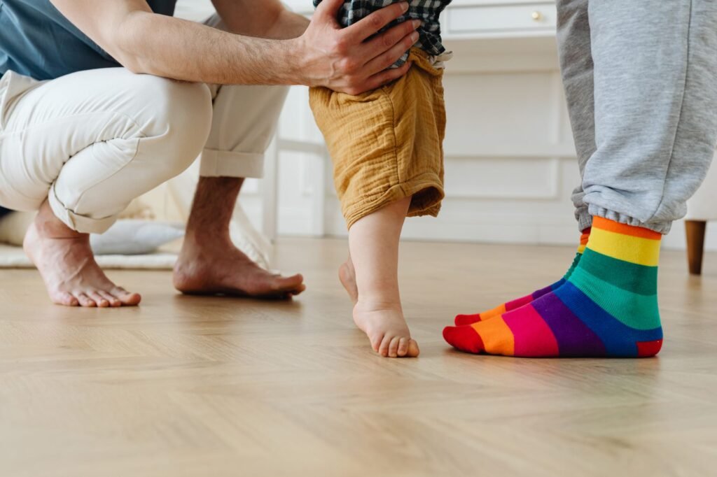 Adult in rainbow striped socks steadying a barefoot toddler taking steps across a wooden floor
