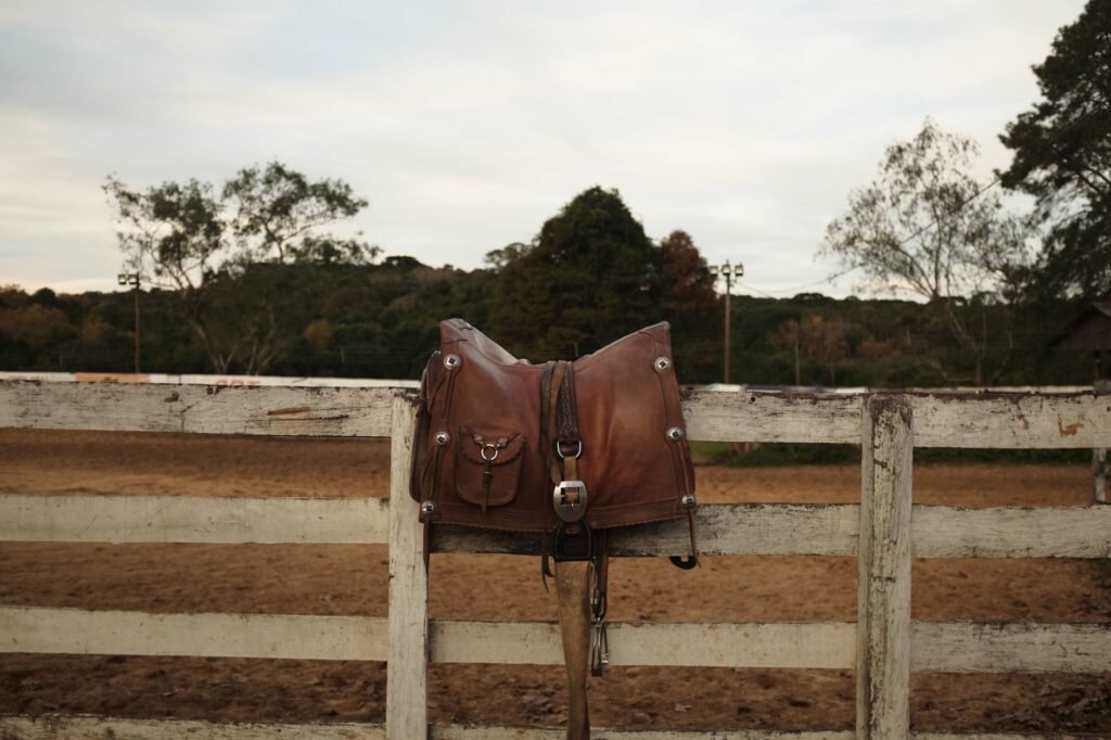 Brown leather saddle resting on a wooden rail fence with trees and a cloudy sky behind