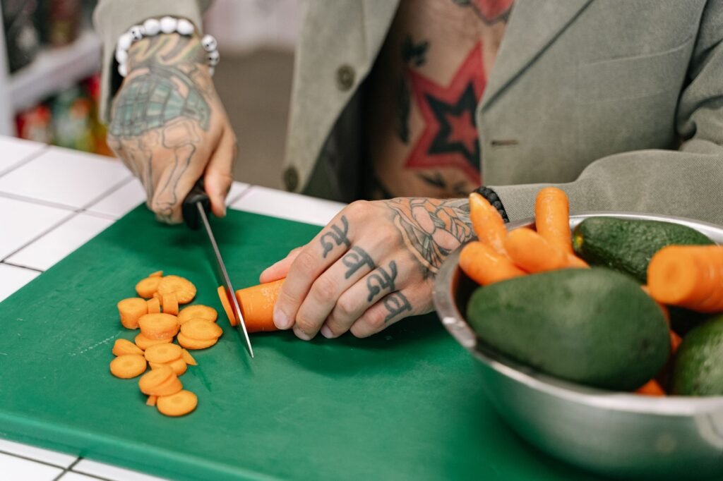 A tattooed person cutting carrots on a green chopping board with avocados nearby, showcasing culinary skill...