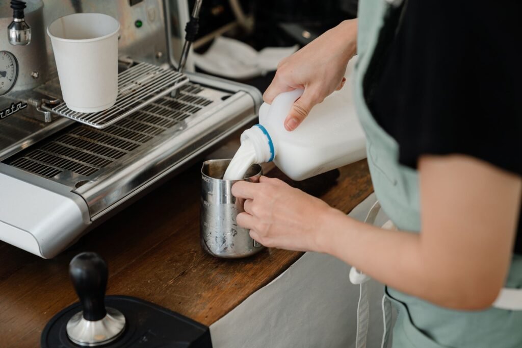 Side view of crop unrecognizable cafe worker pouring fresh milk into big stainless still cup while preparing beverage at counter with coffee machine in modern cafe