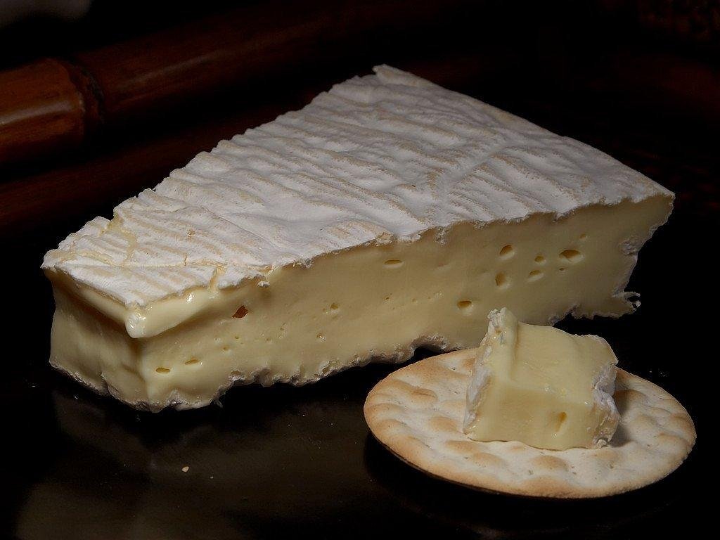 Wedge of soft cheese with a white bloomy rind beside a cracker topped with cheese