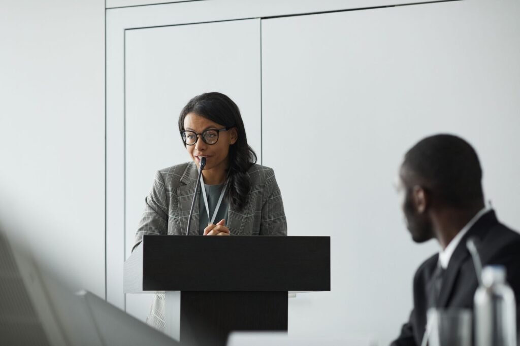 Woman in a gray blazer speaking at a black podium in a bright conference room