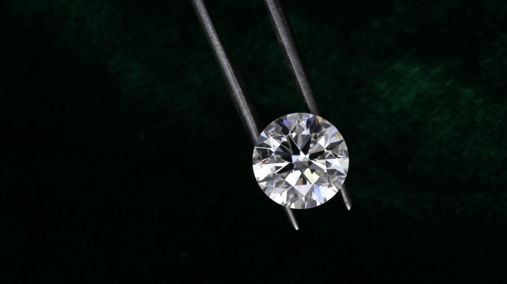 A sparkling round diamond is delicately held by tweezers against a dark textured background