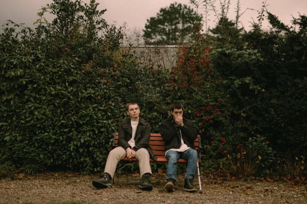 Two men sitting on a bench outdoors in a lush garden setting, surrounded by greenery