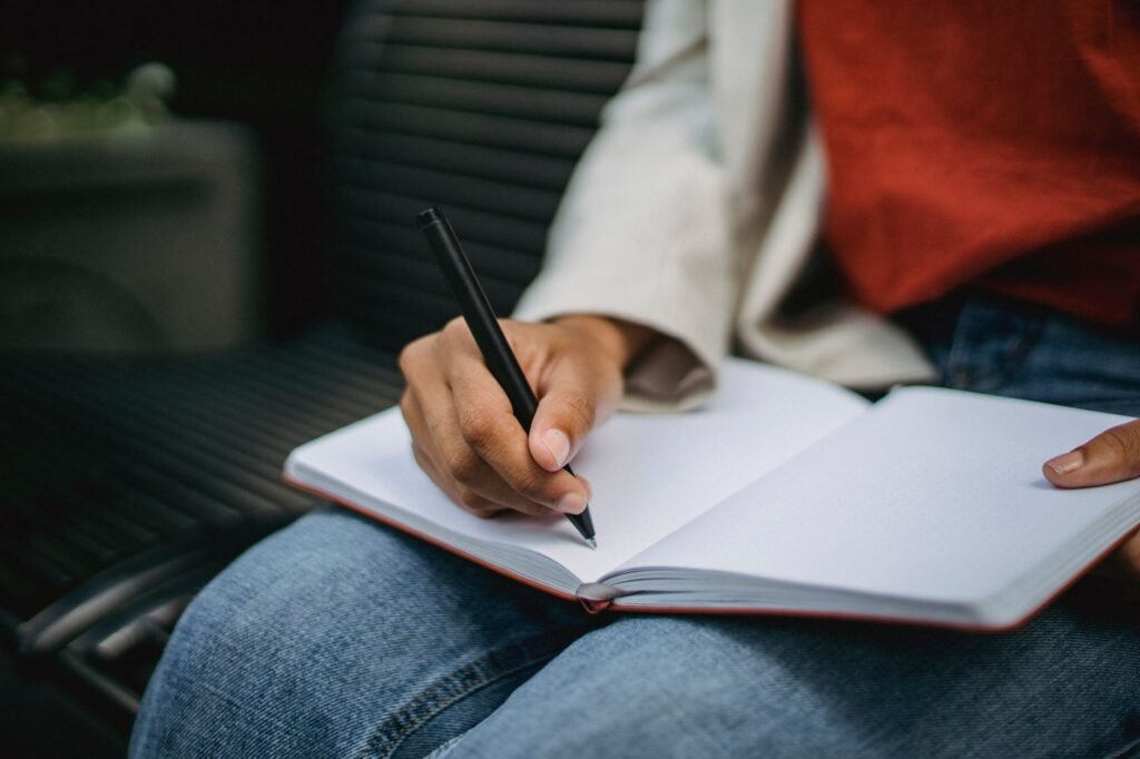 Adult writing notes in a notebook while seated outdoors, pen in hand.