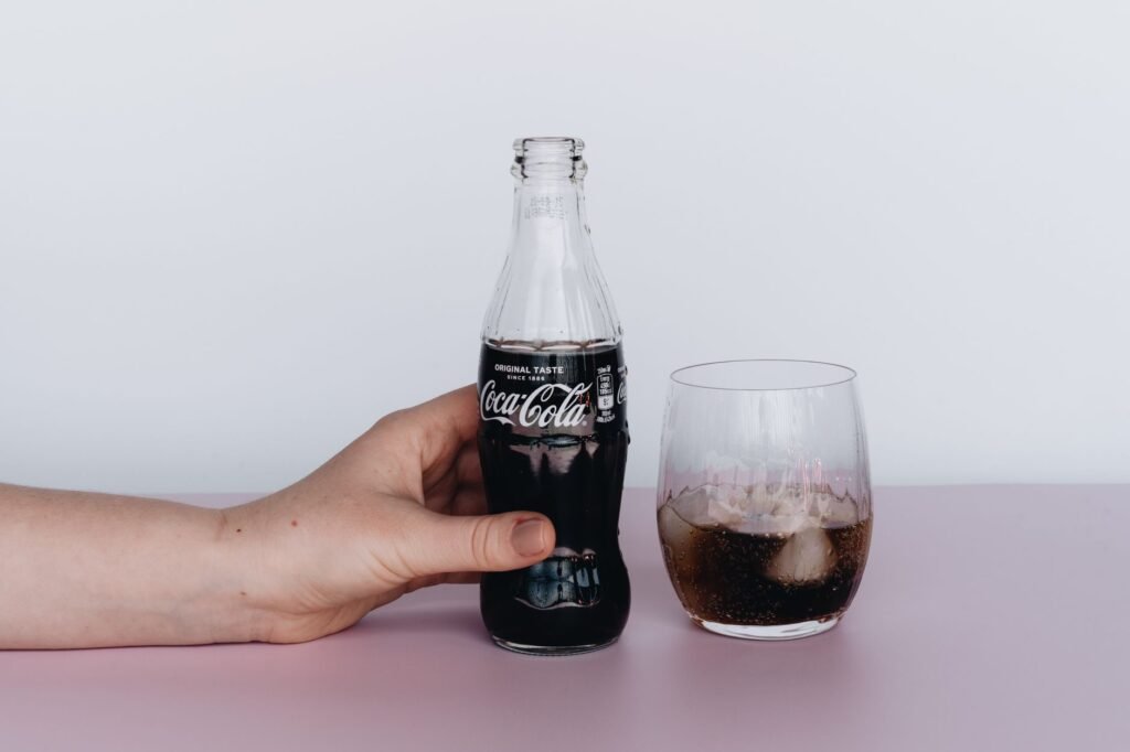 Close-up of a hand holding a Coca-Cola bottle next to a glass filled with ice and soda