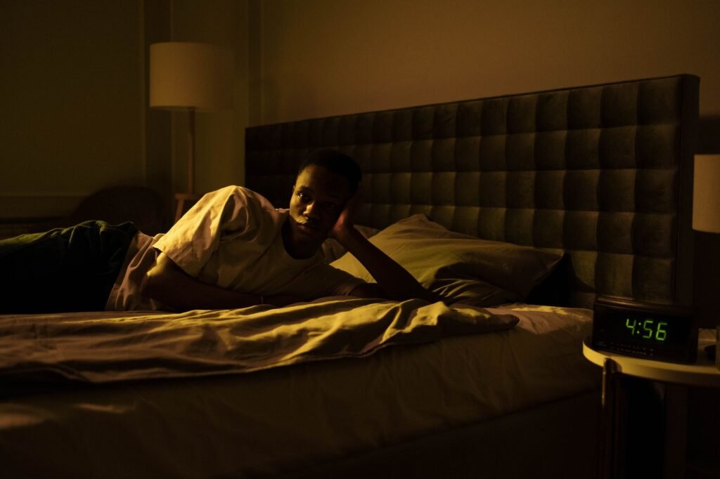 A man lies awake in bed, illuminated by early morning light from a clock reading 4:56 AM.