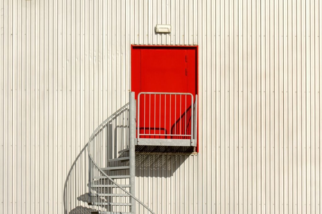 Red door opening onto a railed landing with a metal spiral staircase against a white ribbed wall
