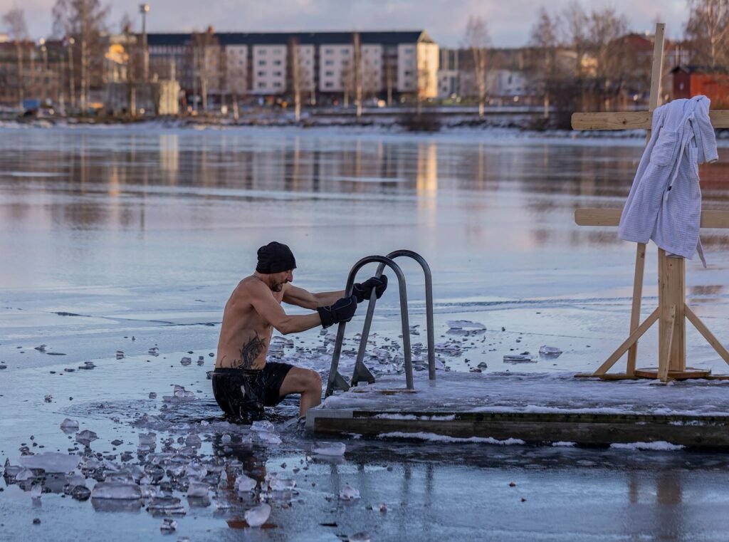 Shirtless man in a knit hat gripping a metal ladder at the edge of an icy lake