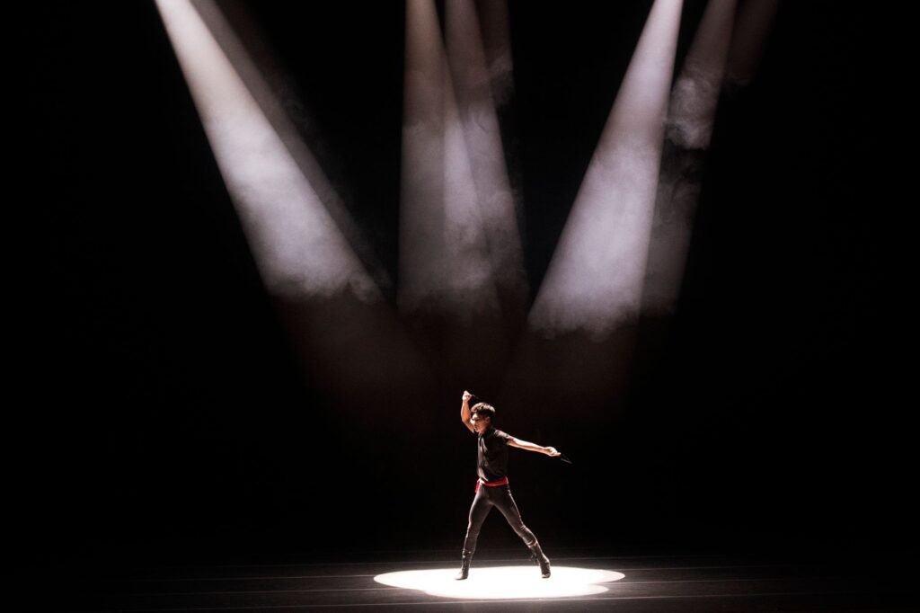 Lone dancer on a dark stage standing in a pool of light under several spotlight beams