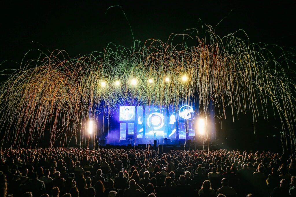 Large crowd facing a brightly lit outdoor stage as streamers fall through the night air