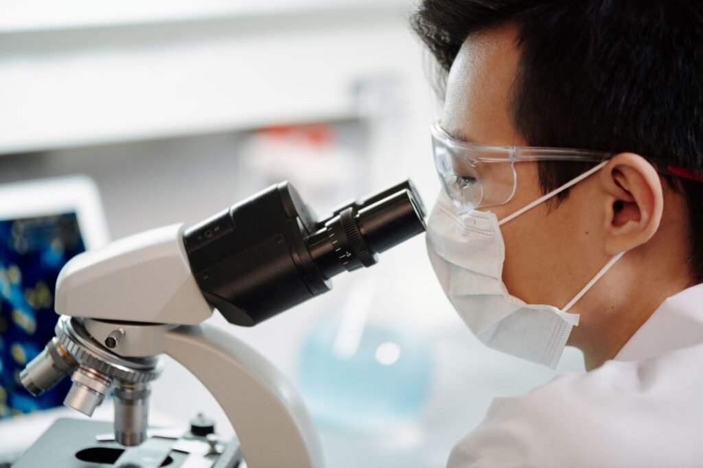 Scientist in safety glasses and a face mask looking into a laboratory microscope