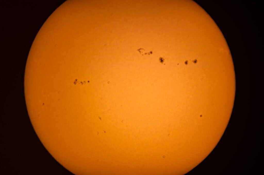 Orange disk of the sun with small dark sunspots against a black sky