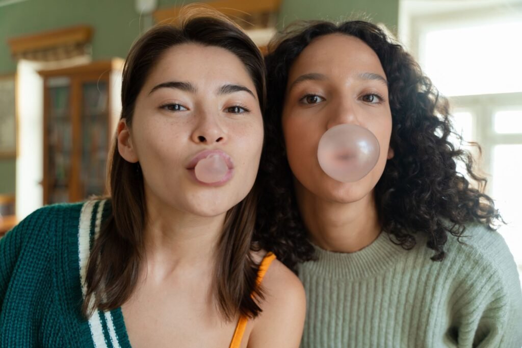 Two young women blowing pink bubble gum bubbles side by side indoors