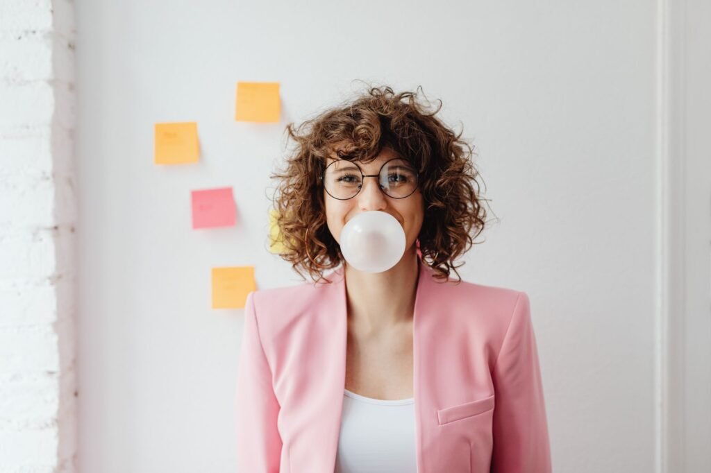 Woman in a pink blazer blowing a large pink bubble gum bubble against a white wall