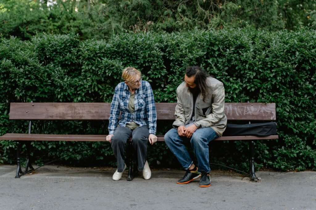 Man and woman sitting apart on a park bench looking down in front of a hedge