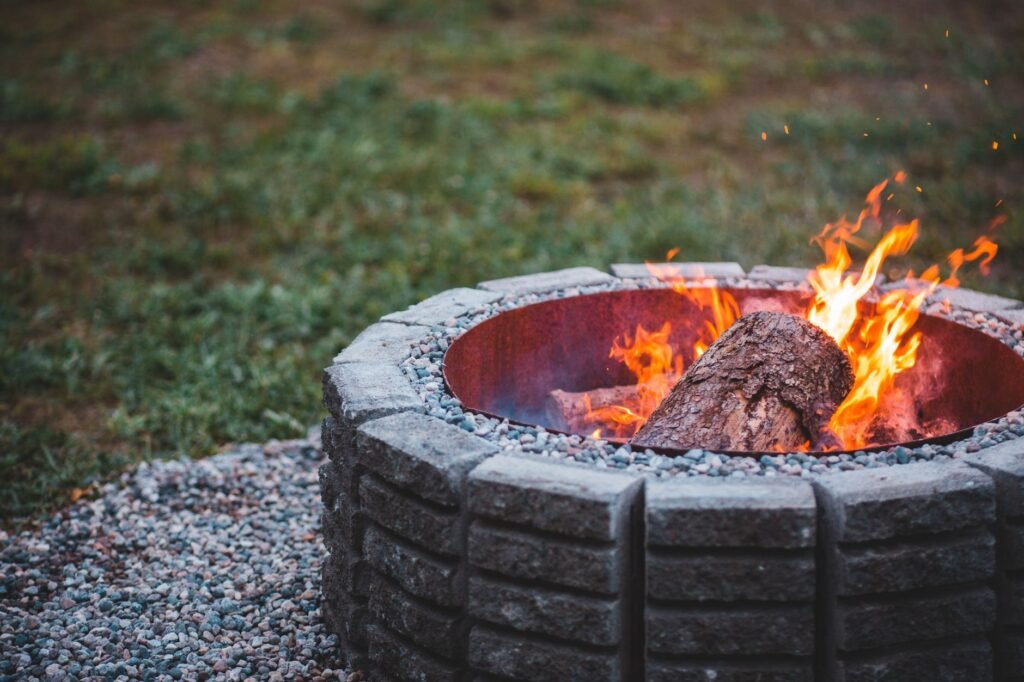 Round stone block fire pit with a crackling log fire on a gravel base in a grassy yard