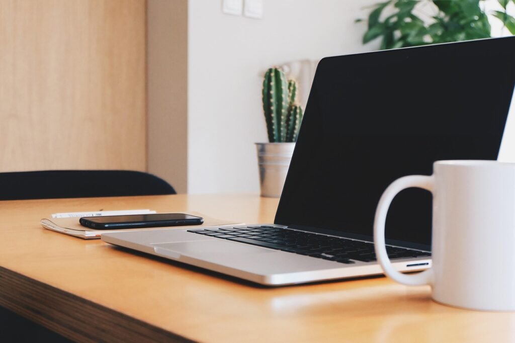 Open laptop, white mug and small cactus on a light wooden desk