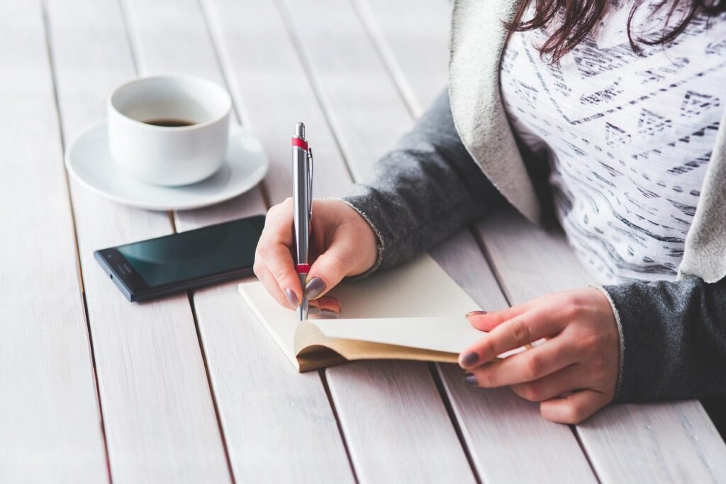 Woman writing in a small notebook at a white wooden table beside a coffee cup and phone