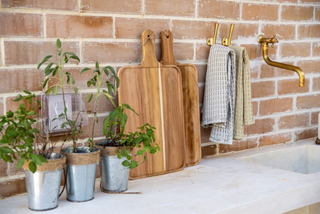 Wooden cutting boards leaning against a brick wall beside potted herbs and dish towels
