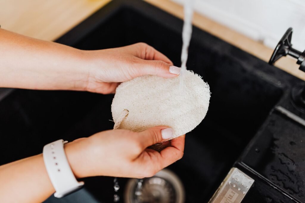 Hands rinsing a natural loofah pad under running water at a dark sink