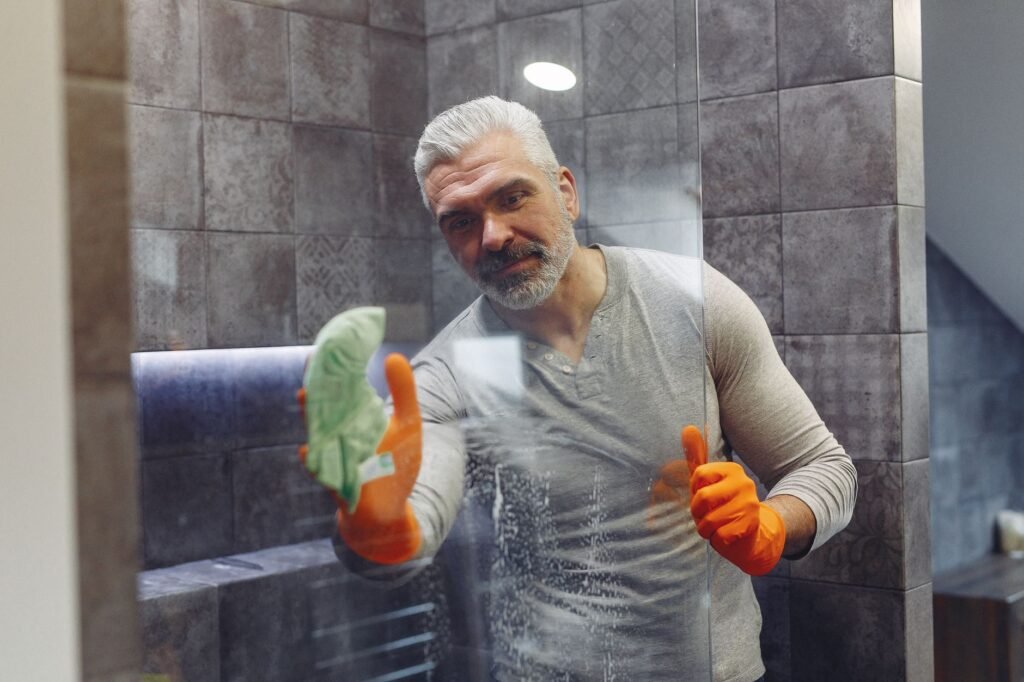 Man in orange rubber gloves wiping a glass shower door with a green cloth