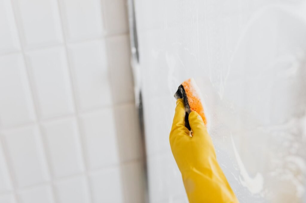 Yellow gloved hand scrubbing white wall tile with an orange sponge