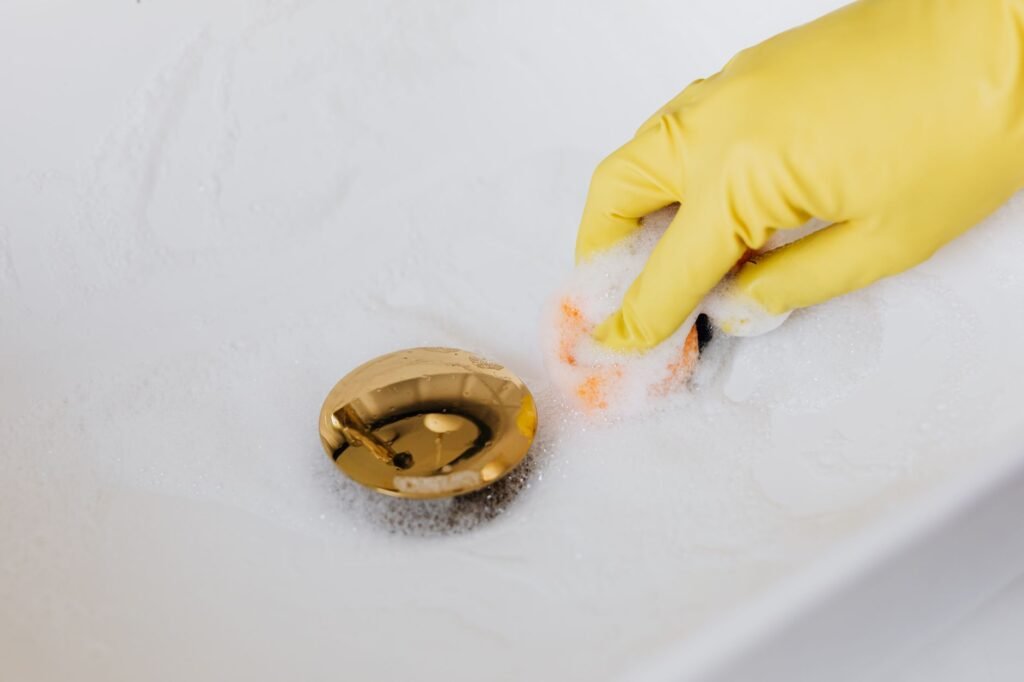 Yellow gloved hand scrubbing a white sink around a gold drain with soapy foam