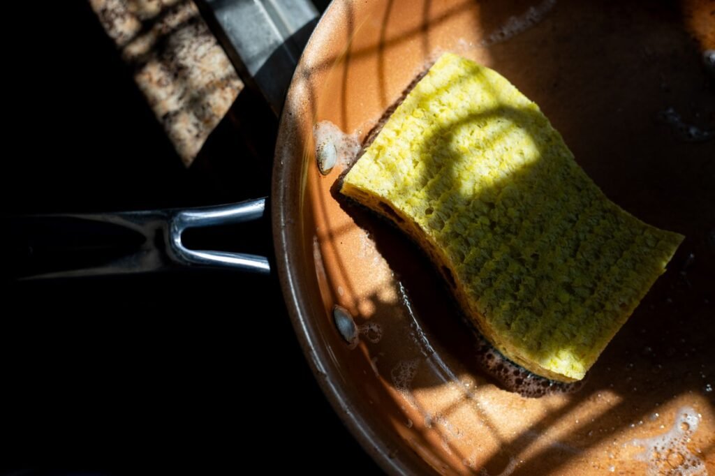 Yellow scrub sponge resting inside a copper frying pan with soapy residue in sunlight