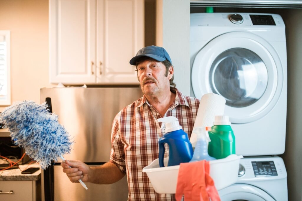 Man holding a duster and a basket of cleaning supplies beside a stacked washer and dryer