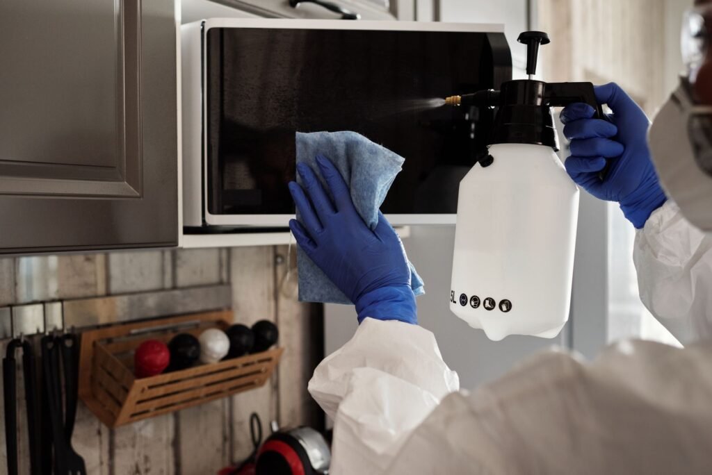 Gloved hands wiping the door of a microwave with a soft blue cloth next to a spray bottle