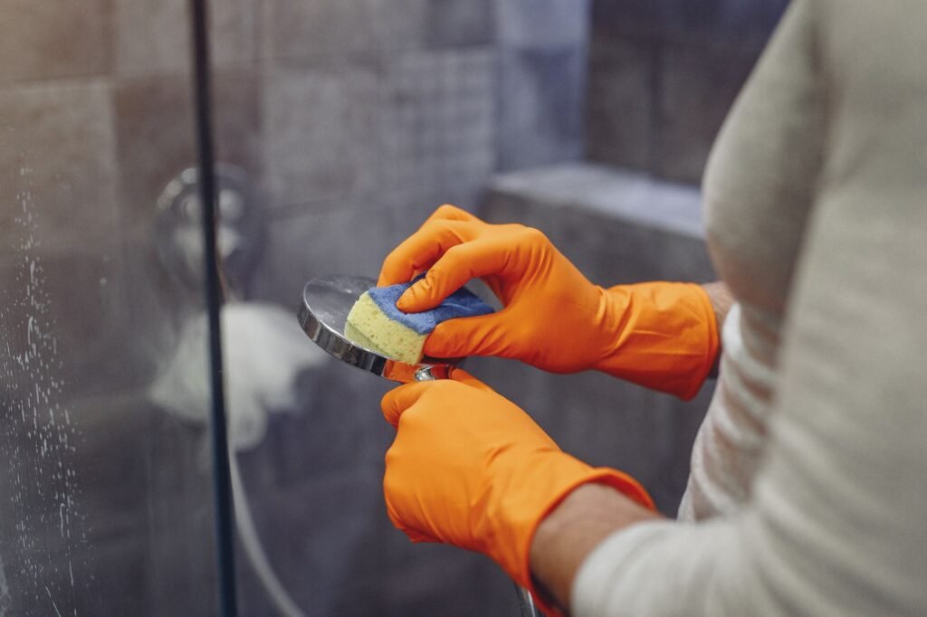 Hands in orange rubber gloves scrubbing a handheld shower head with a sponge inside a shower