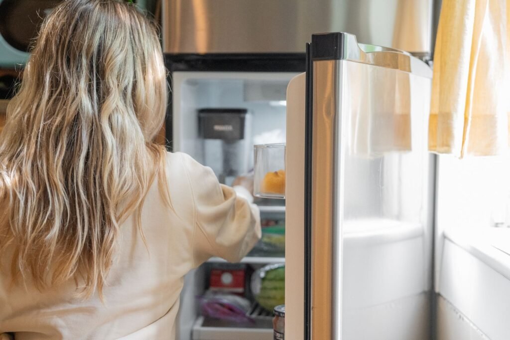 Woman with long blond hair reaching into an open refrigerator stocked with food