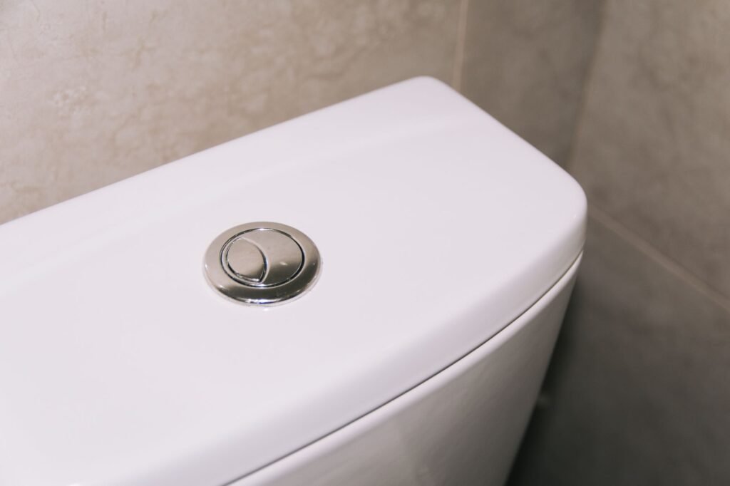 White toilet tank with a round chrome dual flush button against a beige wall