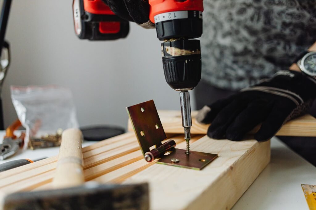 Gloved hands using a cordless drill to drive a screw into a metal hinge on wood