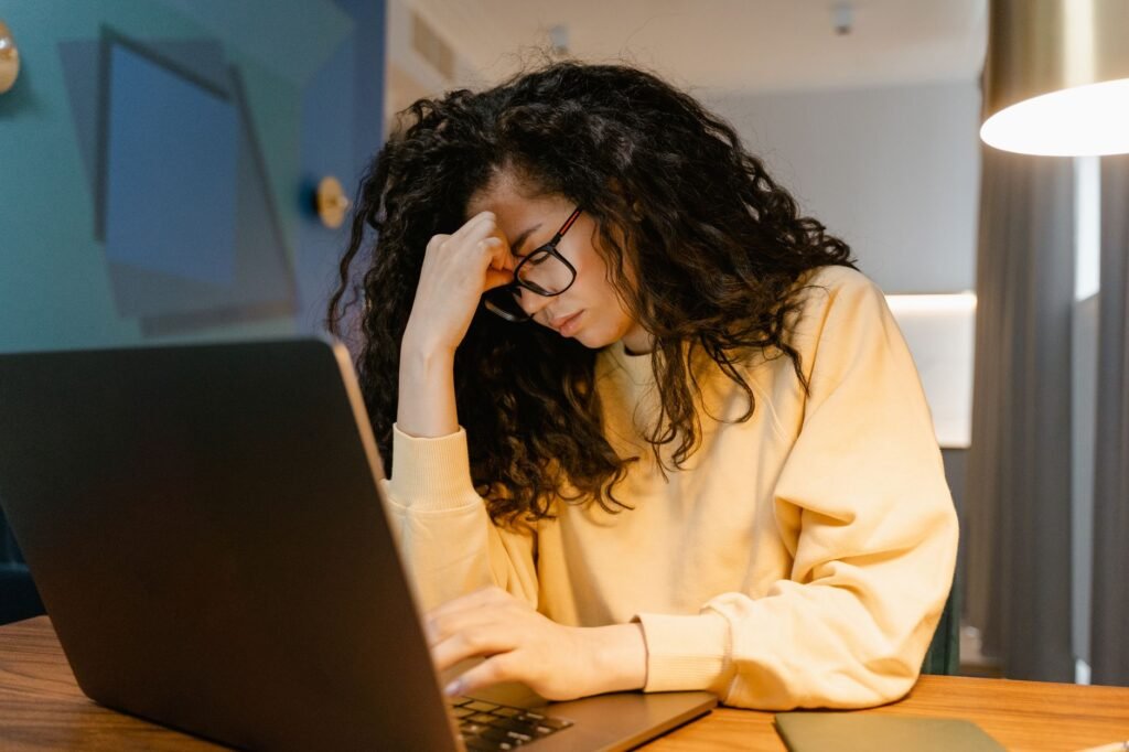 Frustrated woman with curly hair and glasses holding her forehead while working on a laptop