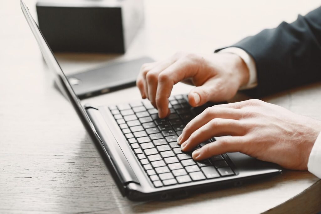 Two hands typing on a black laptop keyboard on a light wooden desk