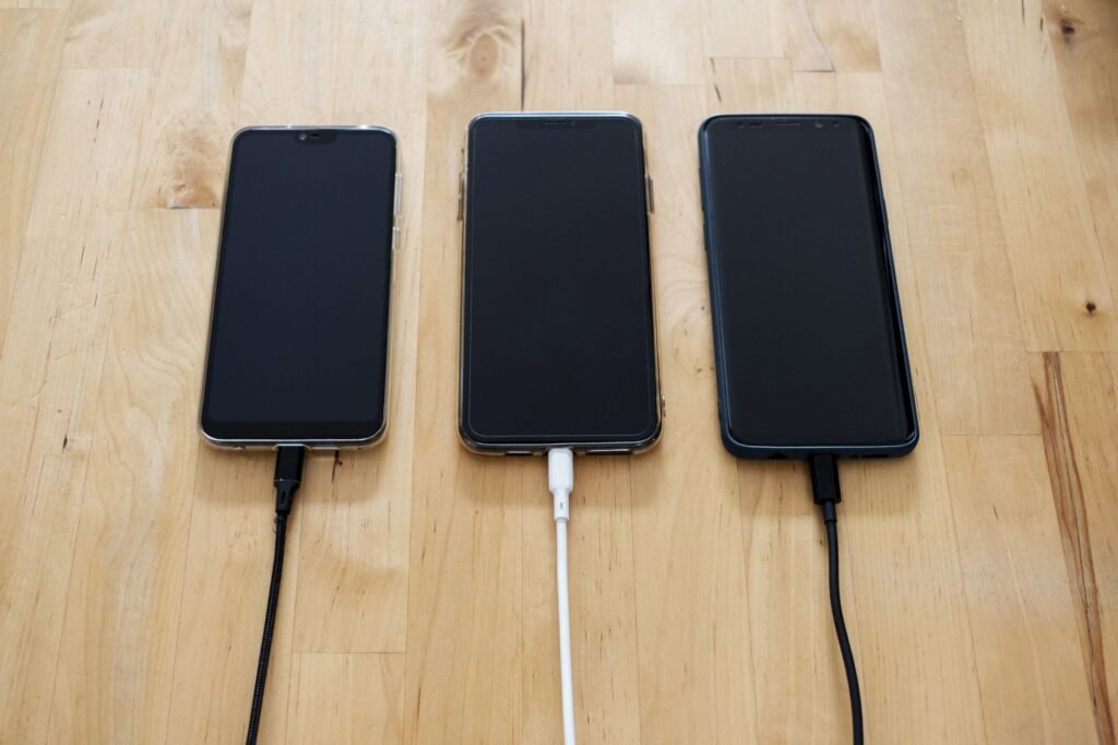 Three smartphones lying face up on a wooden table, each plugged into a charging cable