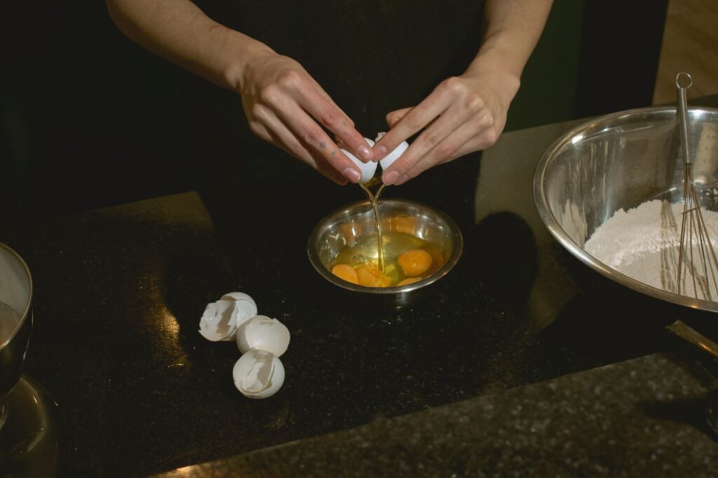 Hands cracking an egg into a small stainless steel bowl of yolks