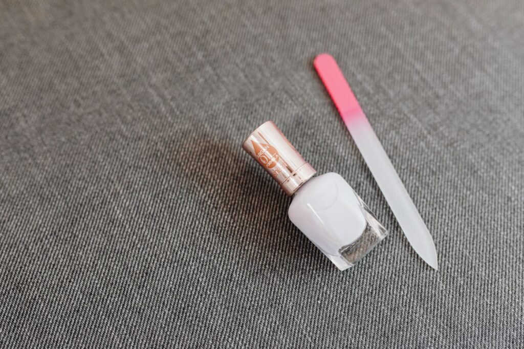 A close-up view of a white nail polish bottle and a pink-tipped nail file on a textured surface, ideal for ...