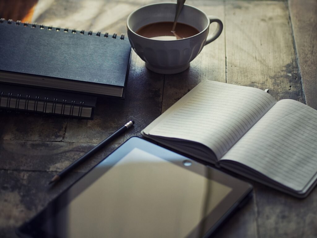 Open notebook, pen, tablet and a cup of coffee on a rustic wooden table