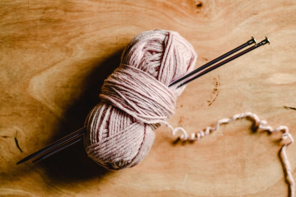 Pink ball of yarn with two metal knitting needles resting on a wooden table