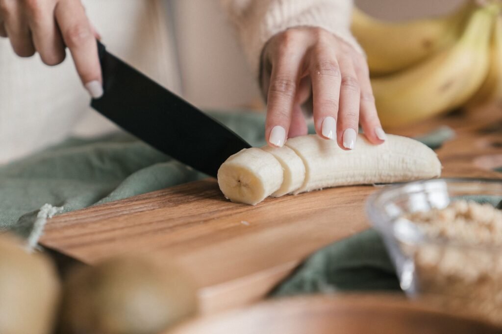 Hands slicing a peeled banana into rounds with a knife on a wooden cutting board