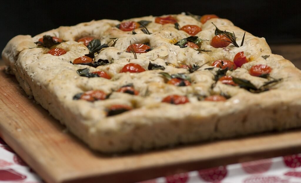 Square focaccia topped with cherry tomatoes and fresh herbs on a wooden cutting board