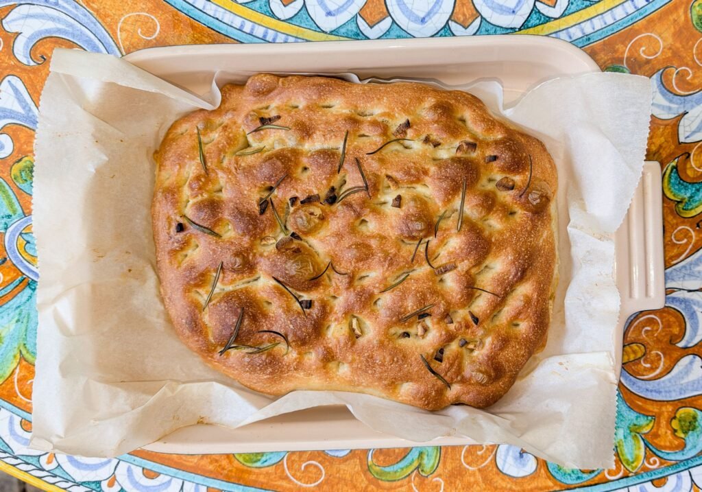 Golden focaccia with rosemary and a dimpled top in a parchment lined baking dish