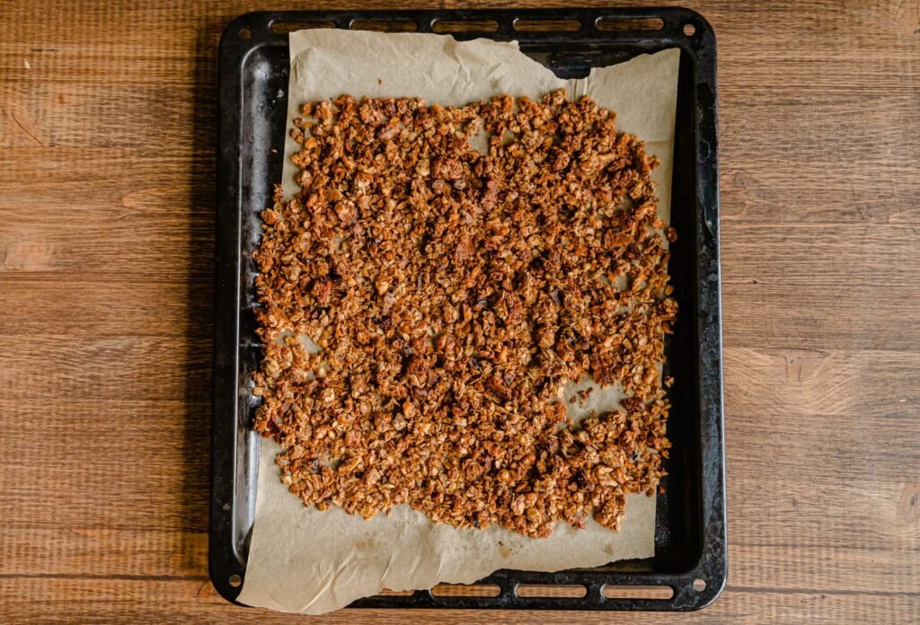 Golden baked granola spread over parchment on a dark baking sheet on a wooden table