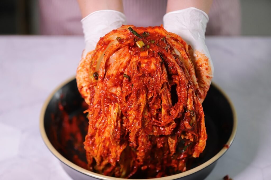 Gloved hands lifting red chili coated napa cabbage leaves over a dark bowl